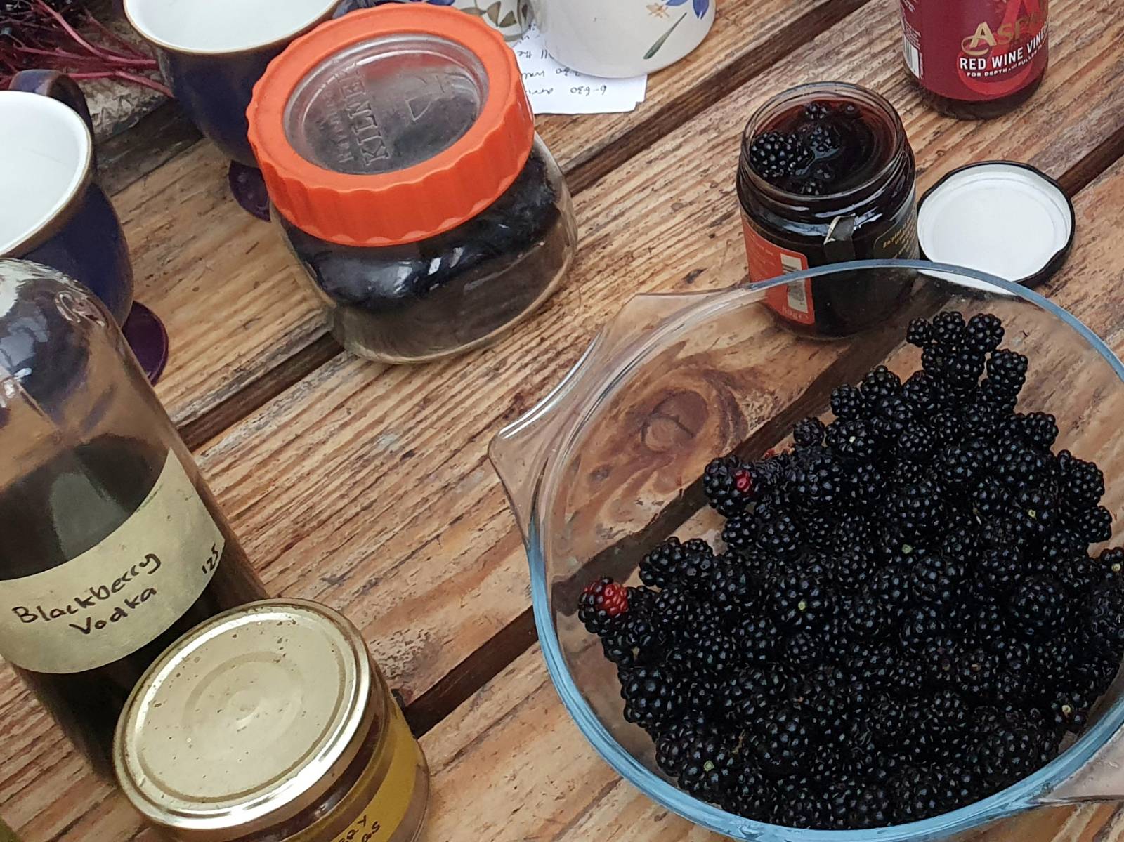 A bowl of blackberries on a wooden table with jars of blackberry vinegar and blackberry powder and a bottle of homemade blackberry vodka. 